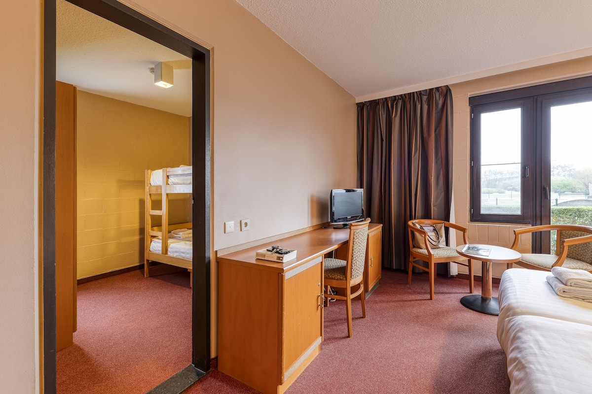 Une chambre d’hôtel avec un lit superposé, un bureau avec une télévision, une chaise, une petite table et un lit.