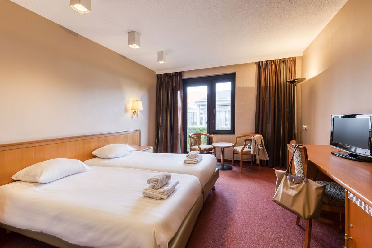 Une chambre d’hôtel avec deux lits, un bureau avec une télévision, une chaise et une fenêtre avec des rideaux.