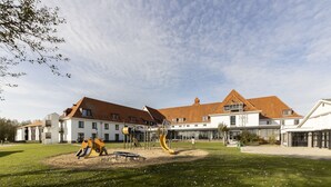 Een speelterrein met glijbanen en klimconstructies, omringd door een verzorgd gazon en een gebouw met een rode pannendak.