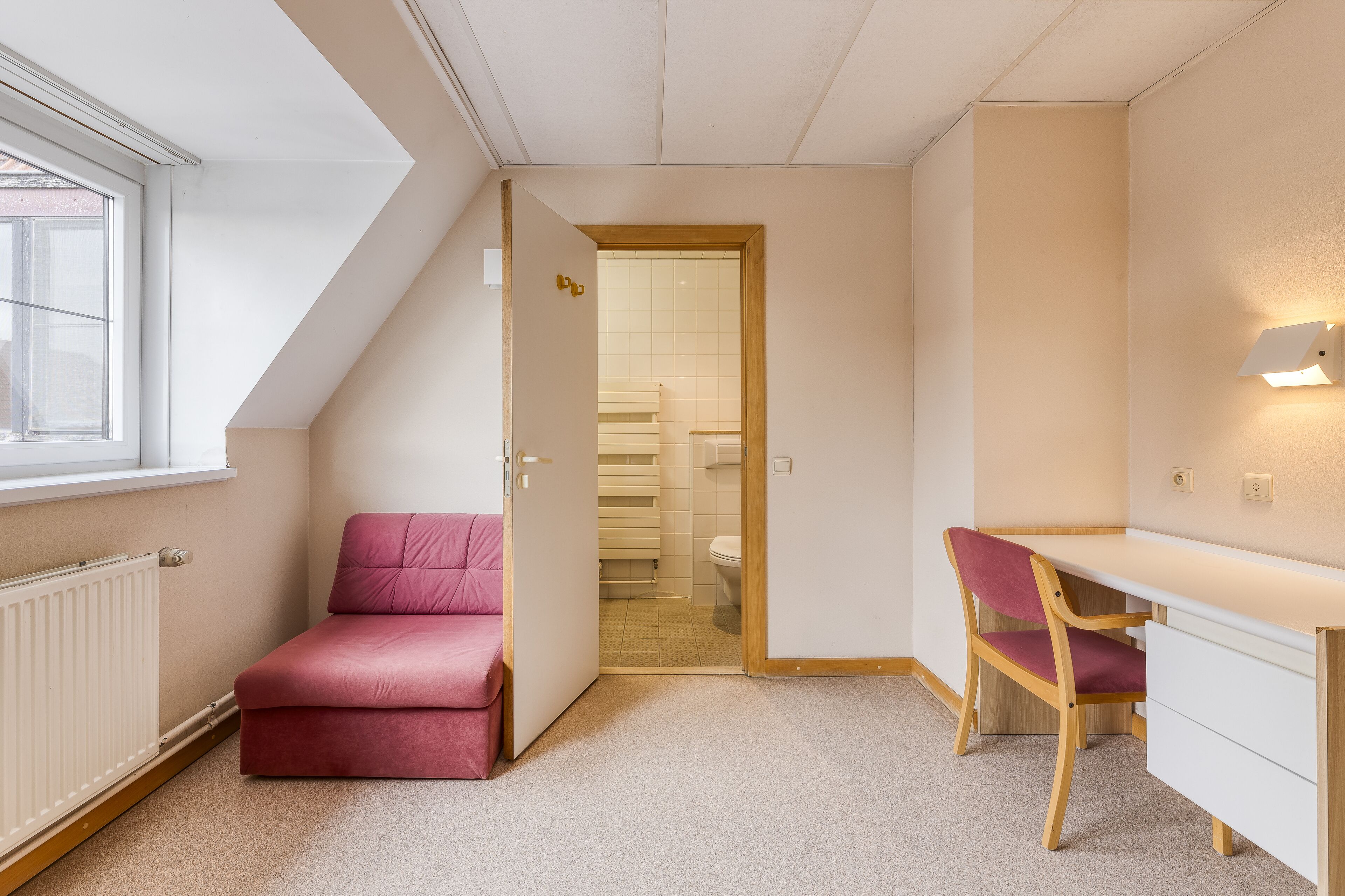 A bedroom with a sloped ceiling, a pink bed, a wooden desk, and a chair.