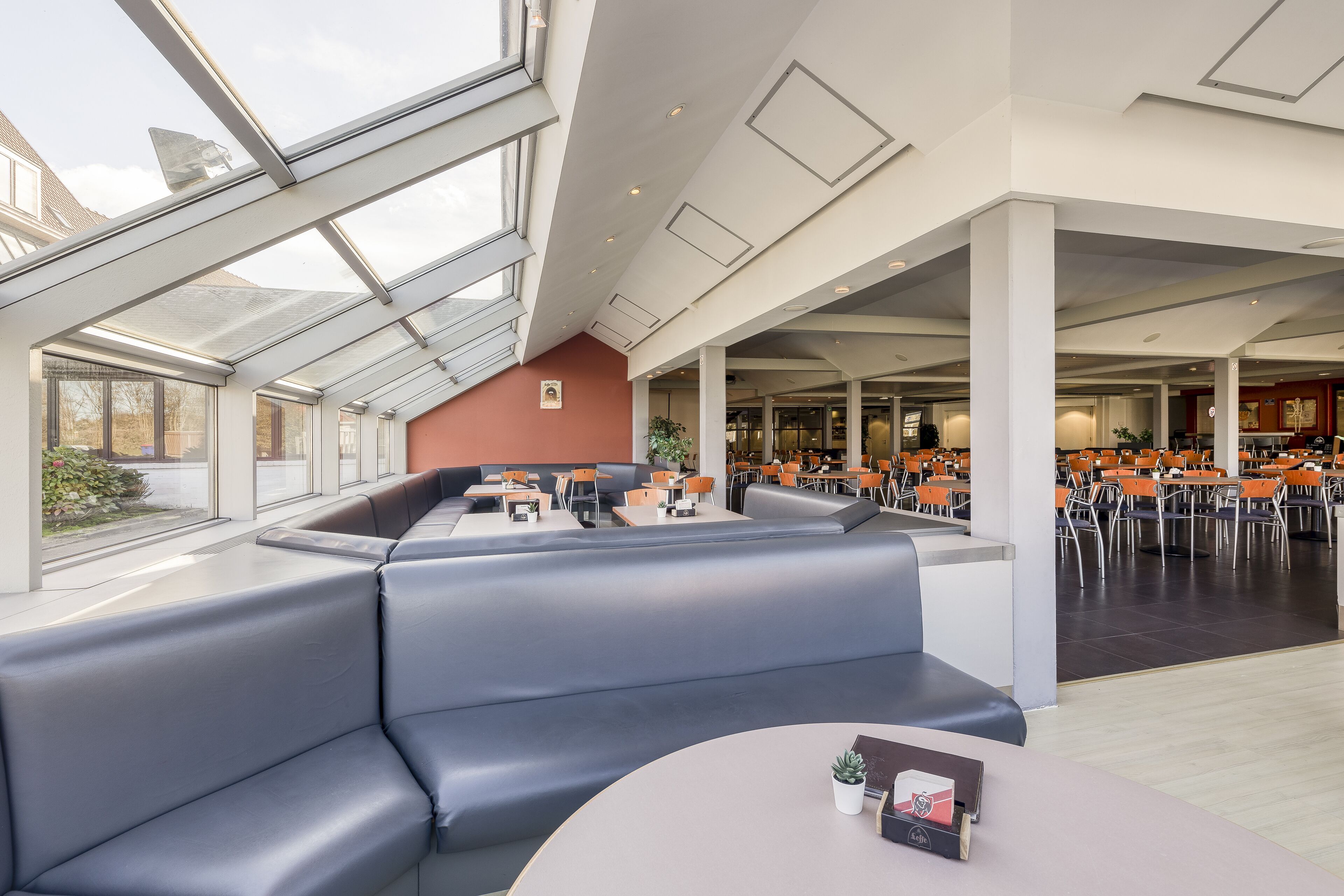 Een modern restaurant met hoge plafonds, grote ramen en een zithoek met blauwe, beklede banken en tafels.