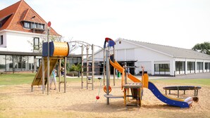 Een speeltuin met diverse speeltoestellen, waaronder een glijbaan en klimrekken, voor een gebouw met een rood dak.