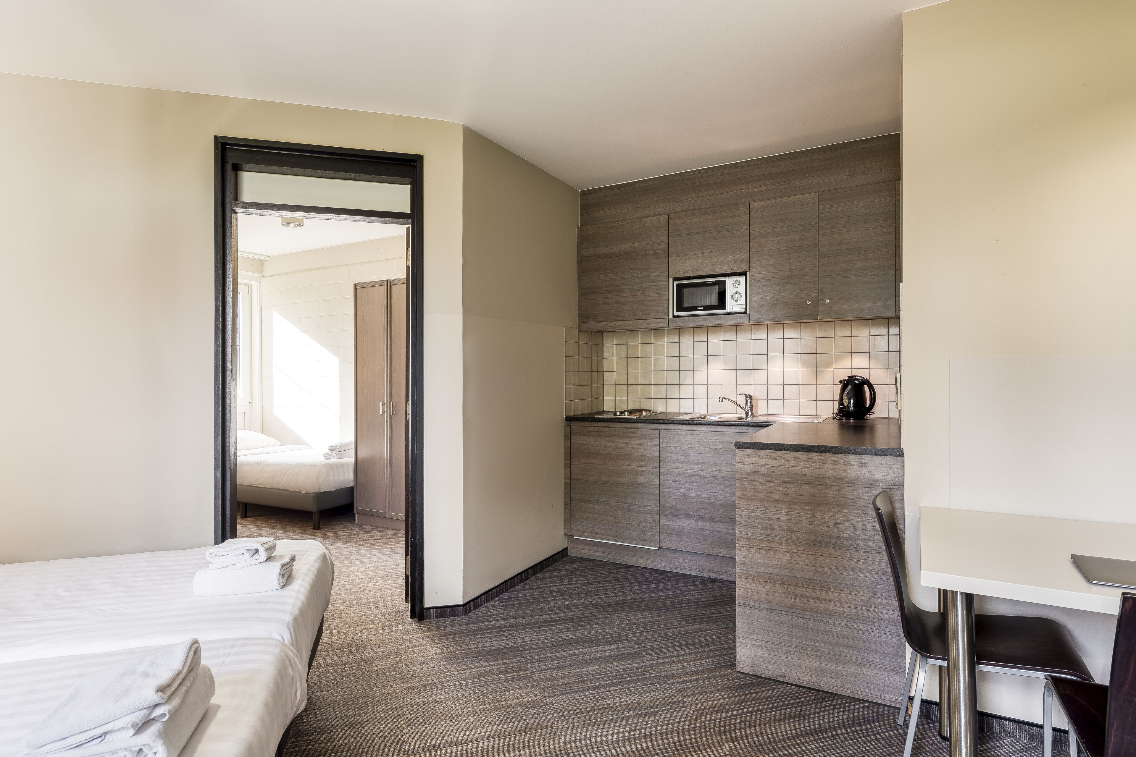 A compact hotel room with a kitchenette, a bed with neatly folded towels, and a view through an open door.