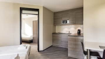 A compact hotel room with a kitchenette, a bed with neatly folded towels, and a view through an open door.