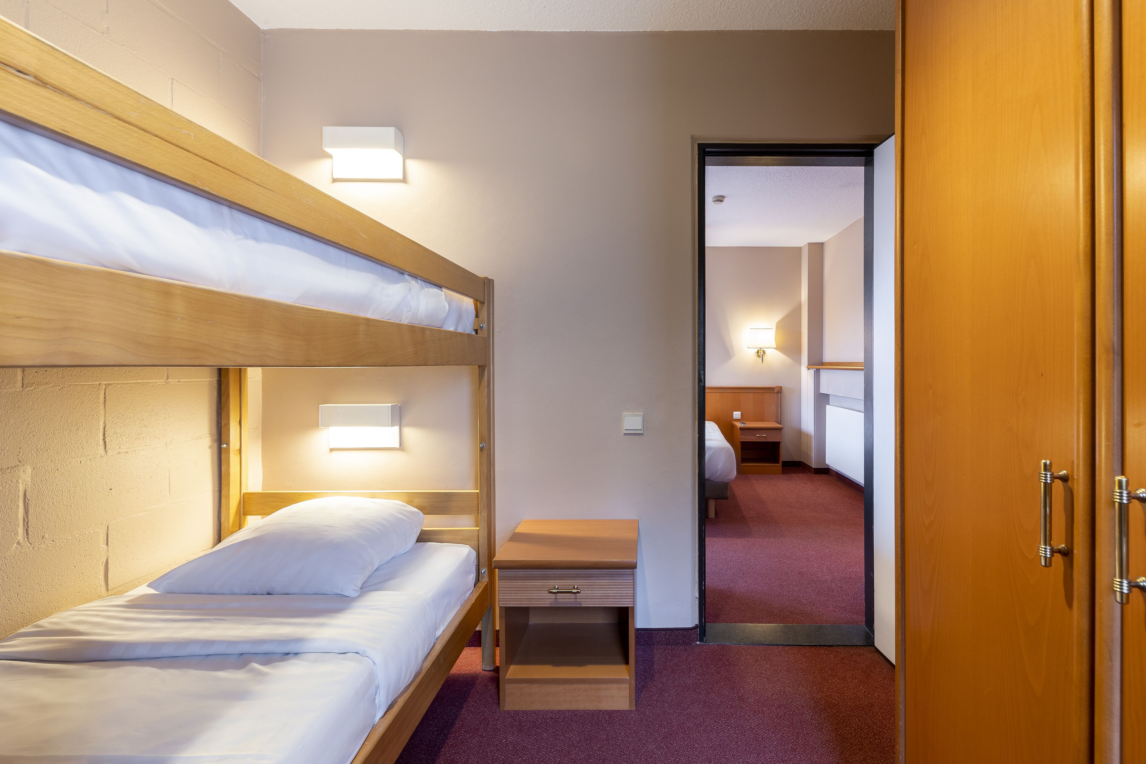 A hotel room with a bunk bed, a bedside table with a lamp, and a wooden wardrobe.