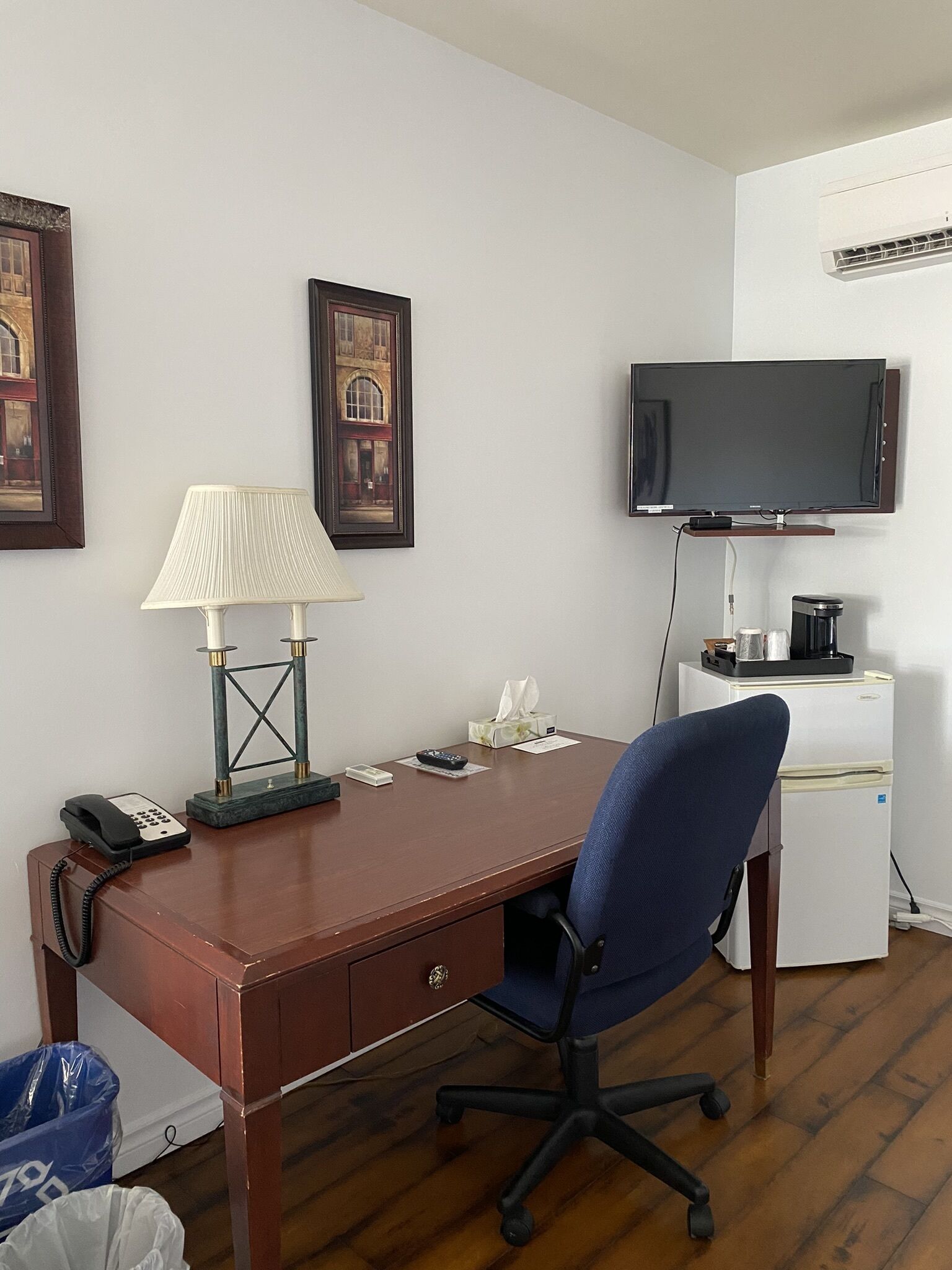 Una habitación de hotel con un escritorio de madera, una silla de oficina azul, un televisor montado, una lámpara y un cubo de basura.