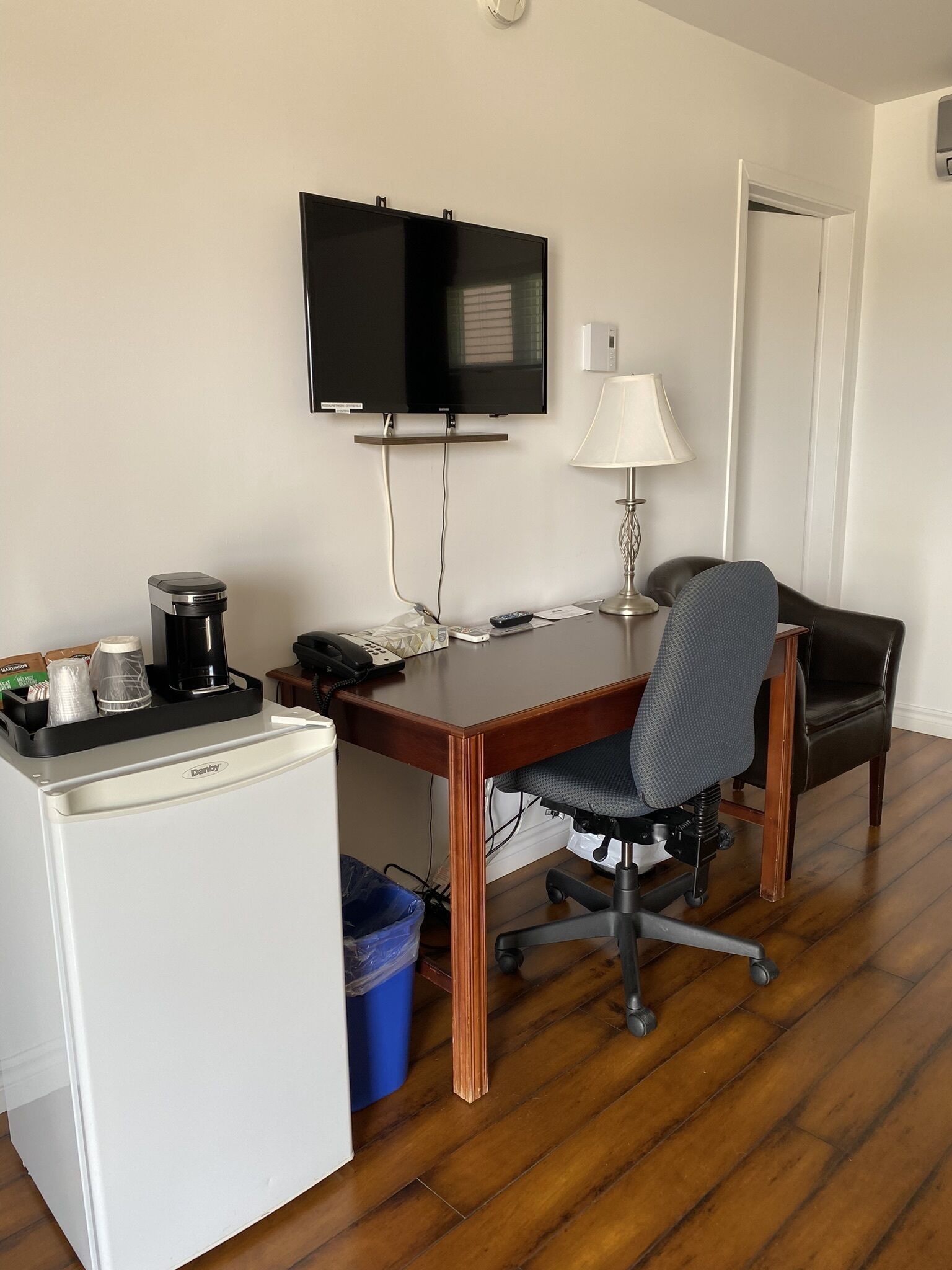 Una habitación de hotel compacta con un escritorio de madera, una silla de oficina negra, un televisor montado en la pared, un refrigerador pequeño y un bote de basura azul.
