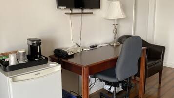 Una habitación de hotel compacta con un escritorio de madera, una silla de oficina negra, un televisor montado en la pared, un refrigerador pequeño y un bote de basura azul.