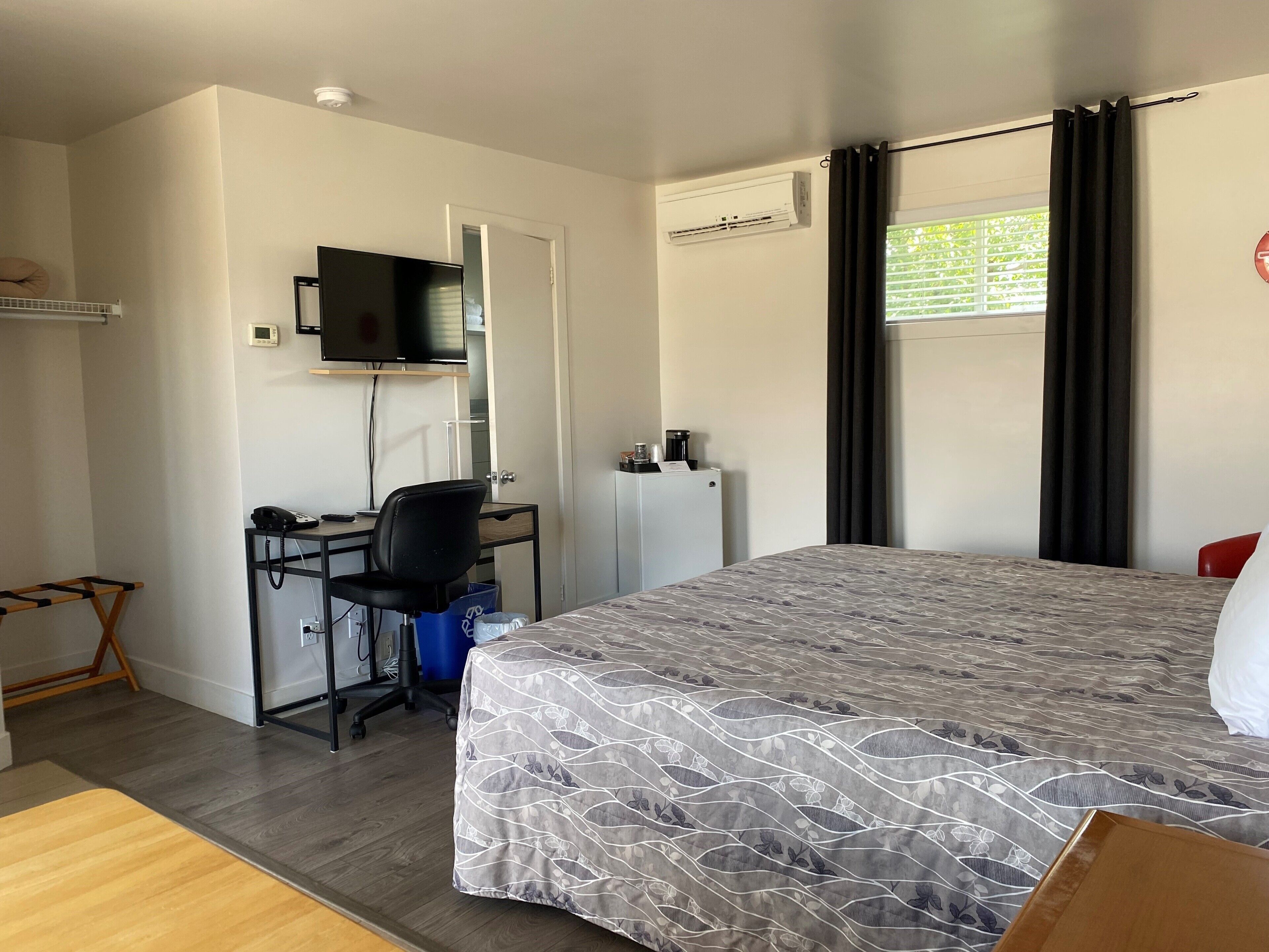 A hotel room with a bed, a desk with a chair, a TV mounted on the wall, an air conditioning unit, and a window with curtains.