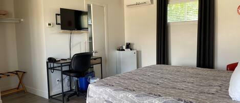A hotel room with a bed, a desk with a chair, a TV mounted on the wall, an air conditioning unit, and a window with curtains.