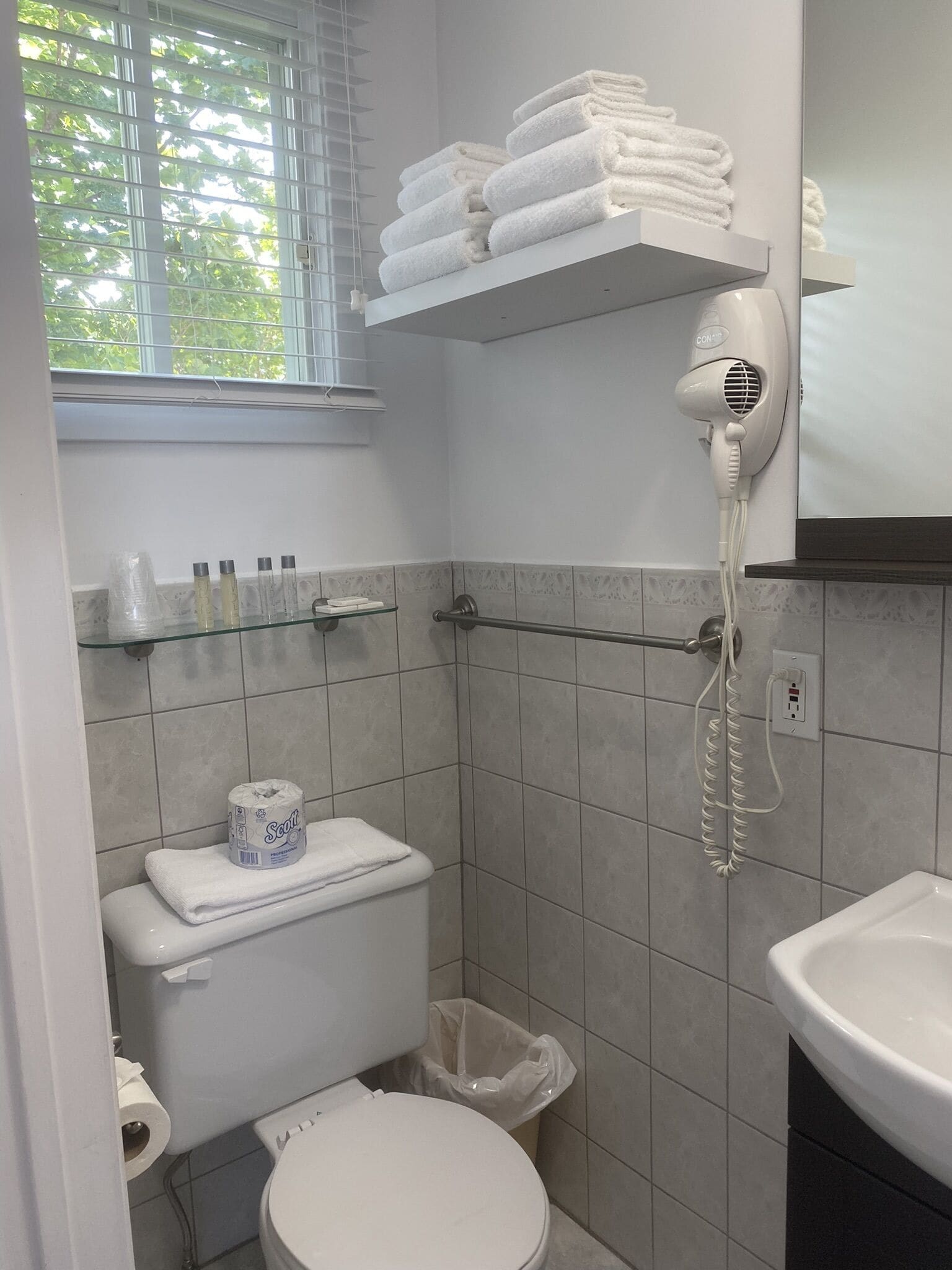 Ein Badezimmer mit Toilette, einem Handtrockner, einer Ablage mit Handtüchern und einem Fenster mit Jalousien.