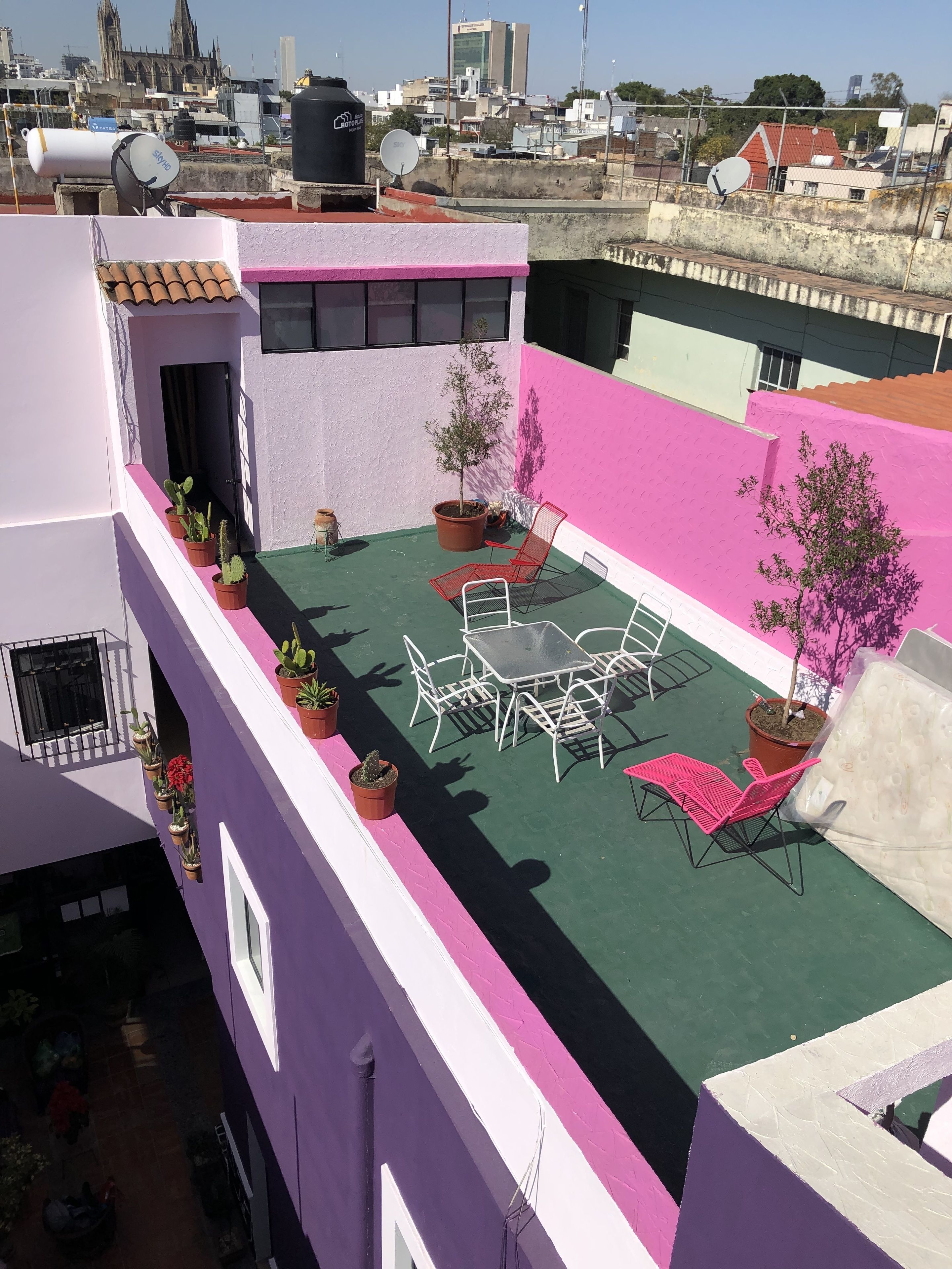 A rooftop patio with a table and chairs, surrounded by potted plants and a pink wall.