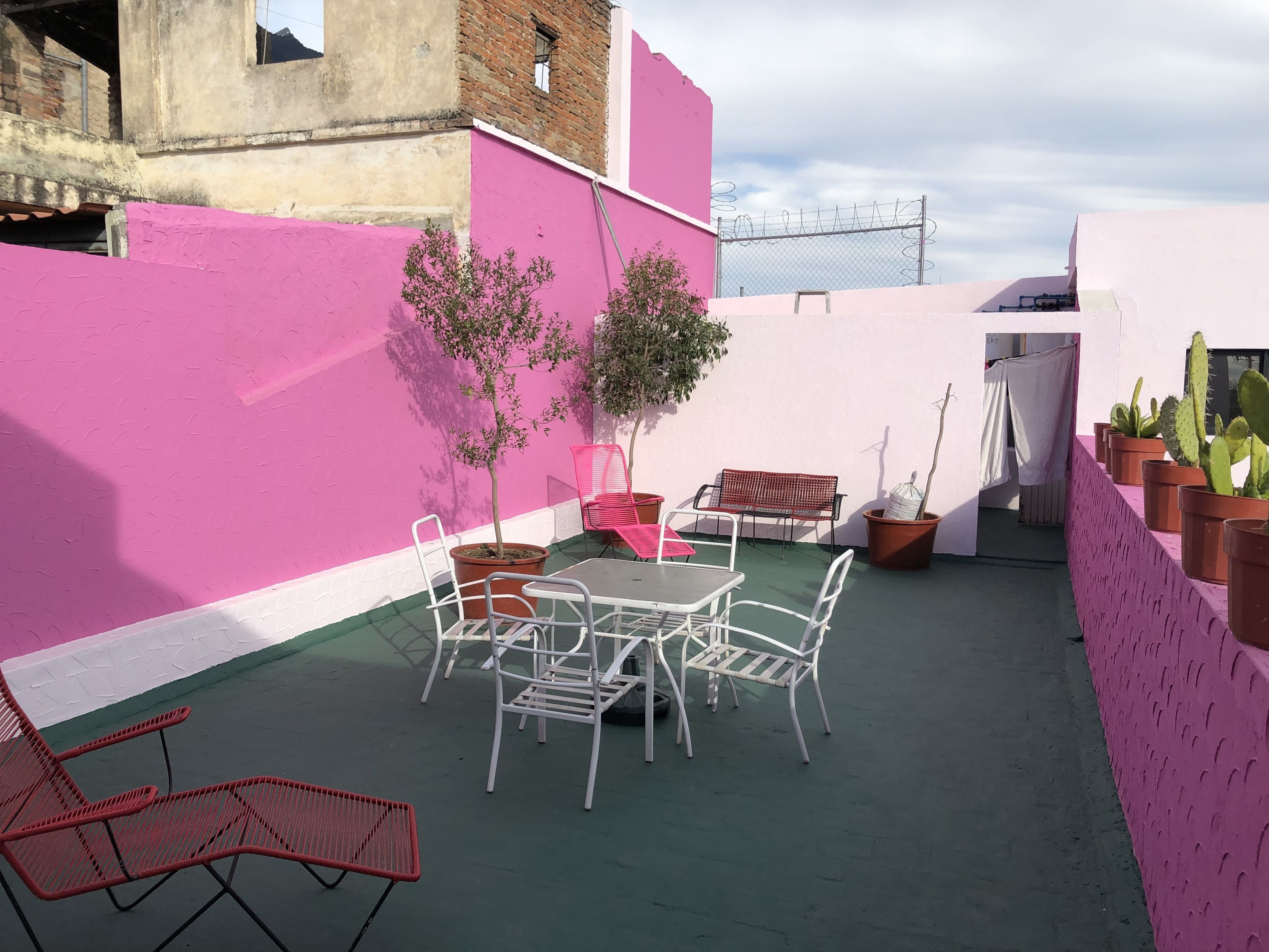 Un terrazzo sul tetto con un muro rosa, un tavolo con sedie, una sedia rossa, piante in vaso e una vetrina di cactus.