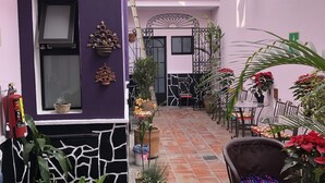 Un patio con pared morada, suelo de baldosas, plantas en macetas, un extintor y mobiliario exterior.