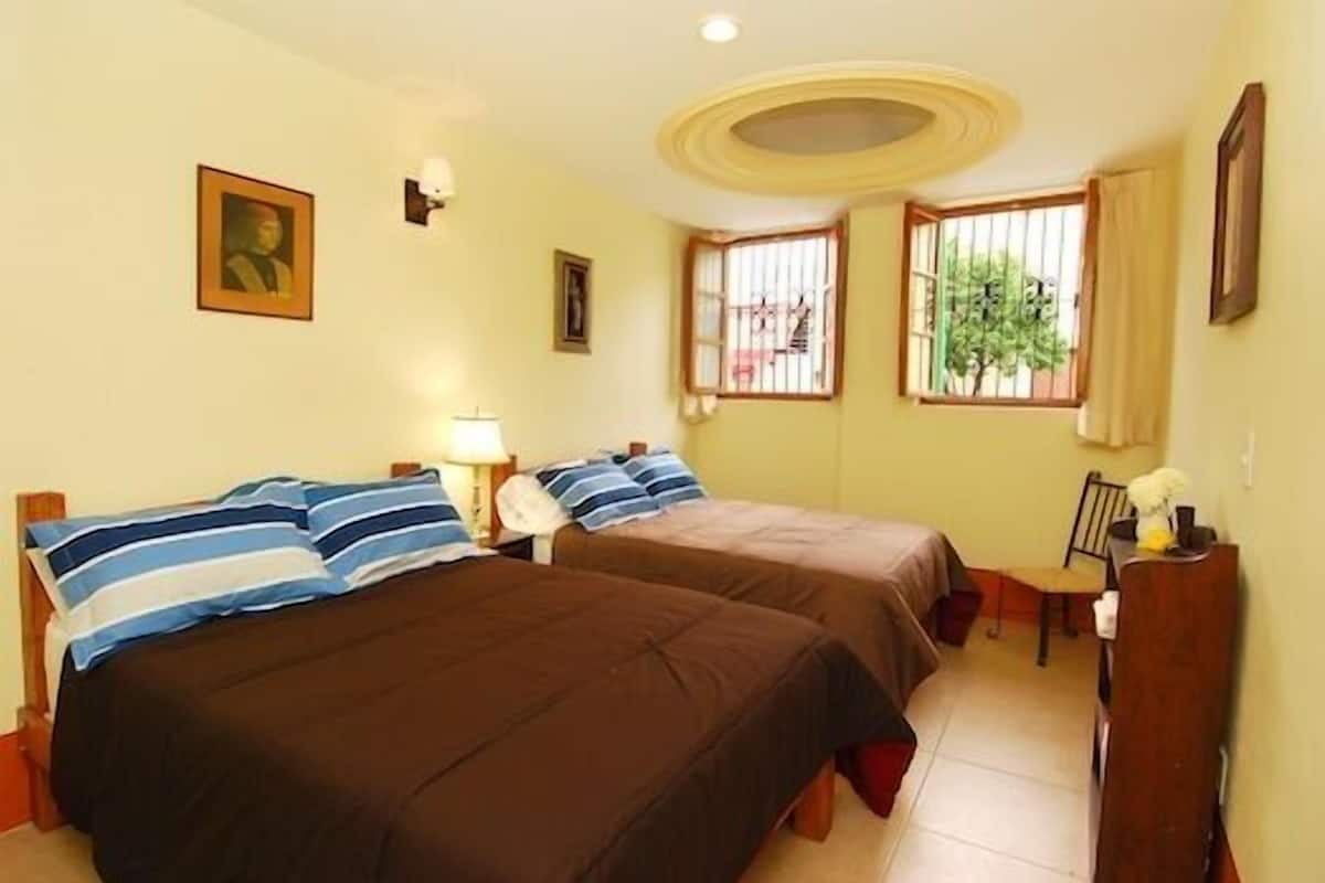 Quarto com duas camas, uma escrivaninha e uma cadeira. Há duas janelas com grades, uma delas com vista para o verde lá fora.