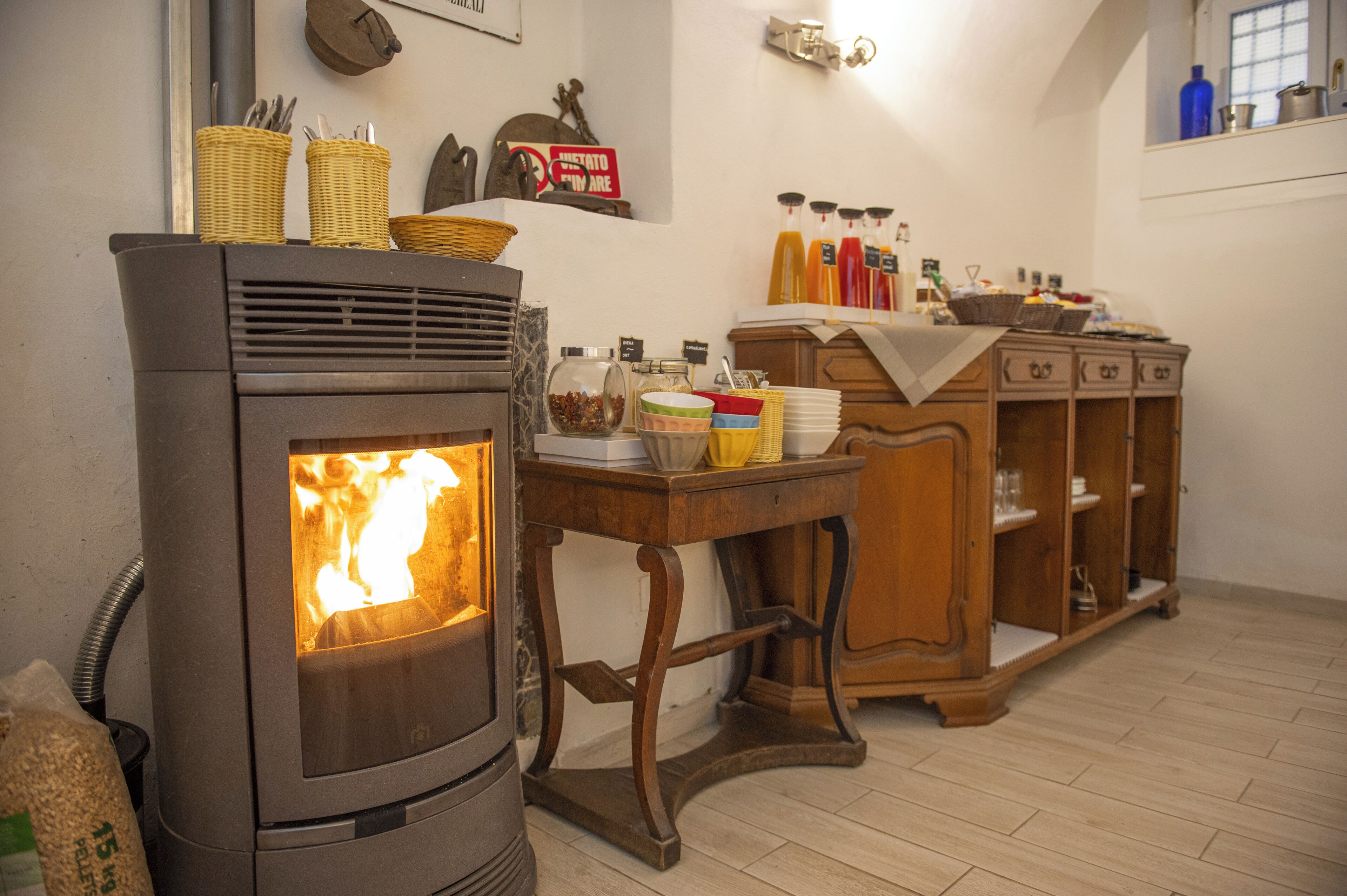 A wood-burning stove with a fire, a wooden table with various items, and a wooden cabinet with shelves.