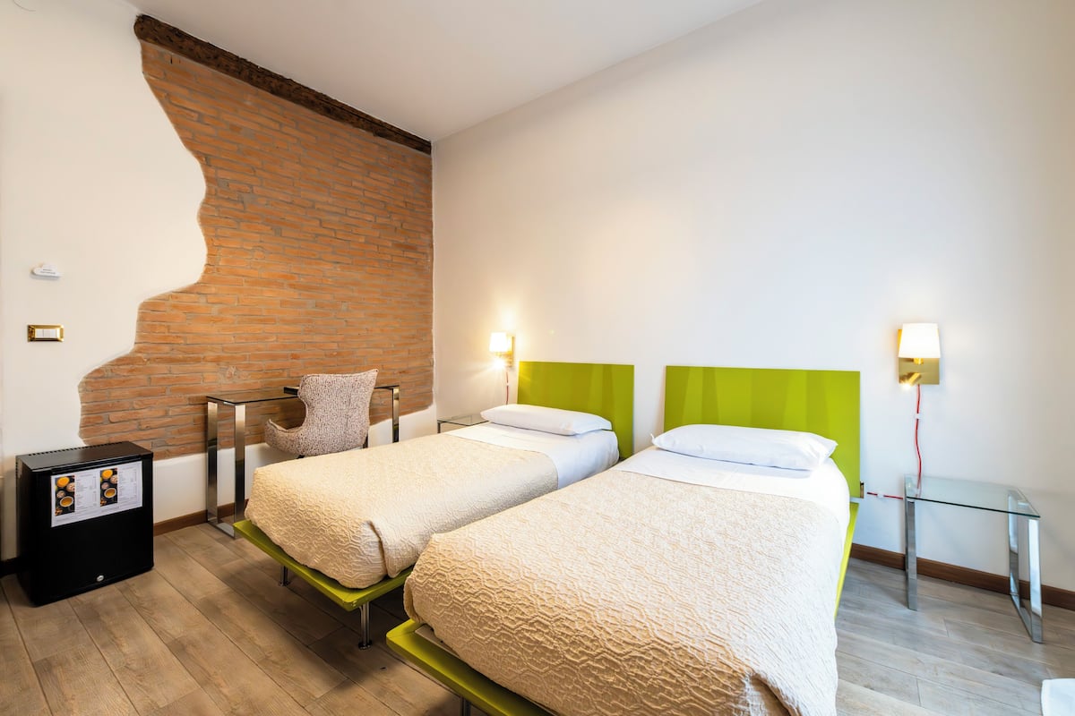 A hotel room with two beds, a brick wall, a small table, and a wall-mounted lamp.