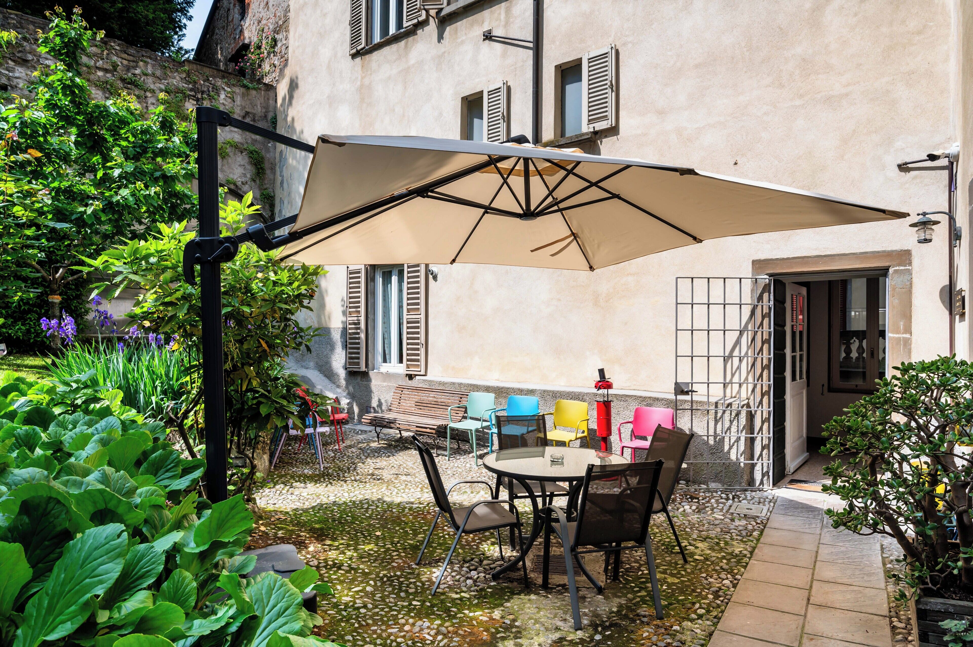 Une terrasse extérieure avec une table et des chaises, entourée de verdure et bordée d’un bâtiment aux volets fermés.