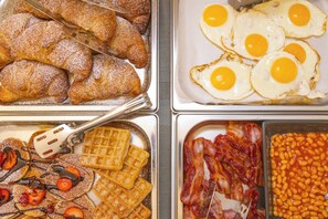 A breakfast buffet with pastries, fried eggs, waffles, bacon, and beans.