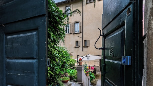 A narrow alleyway with a black door on the left and a black door on the right, leading to a building with a beige facade and shutters. - Palazzo Rivola (Bergamo)