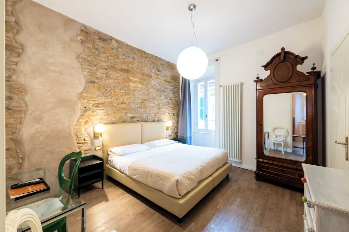 A bedroom with a stone wall, a bed, a dresser, a mirror, and a window with curtains.