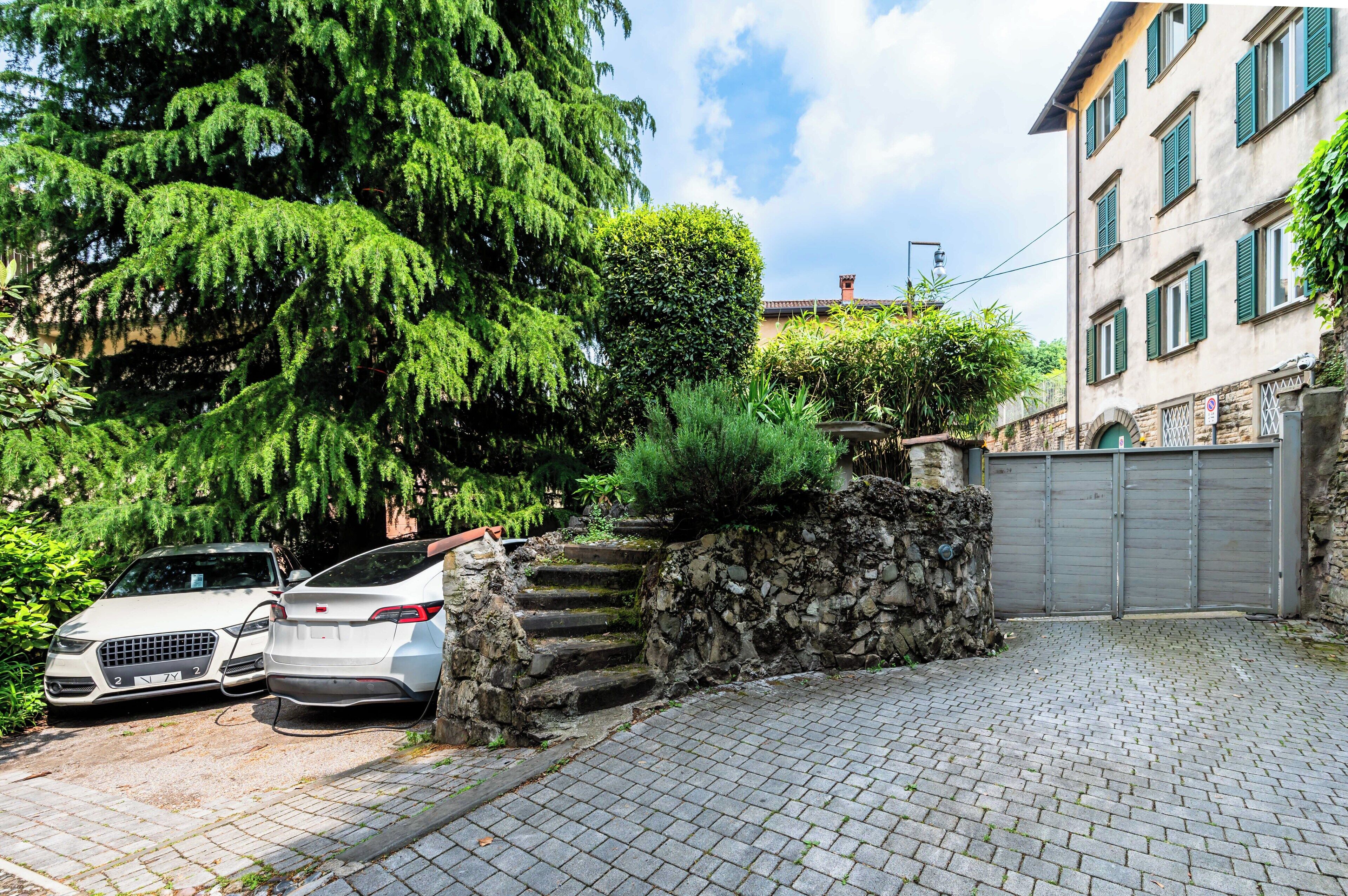 Uma entrada de automóveis pavimentada que leva a um muro de pedra com degraus, dois carros estacionados e um edifício com persianas verdes.