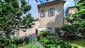 A courtyard with a building, lush greenery, and a pathway. - Palazzo Rivola (Bergamo)