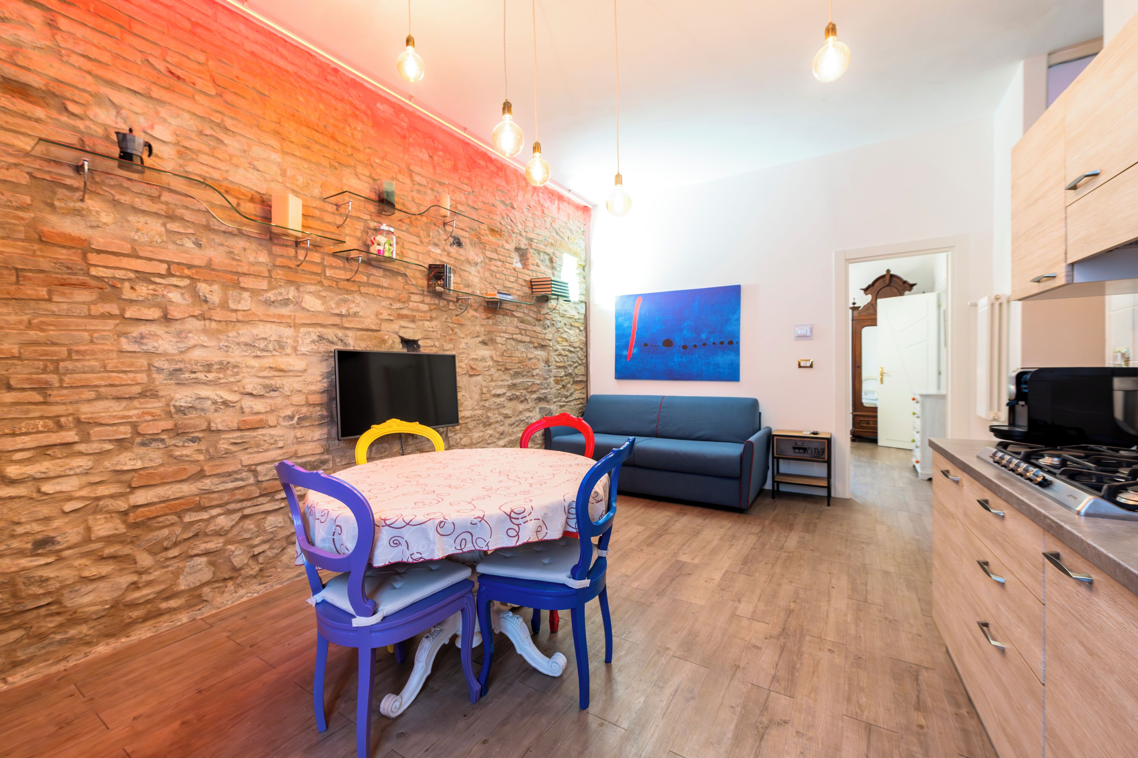 Un coin repas avec une table et des chaises, un mur en pierre, un canapé bleu et une cuisine équipée d’une cuisinière.