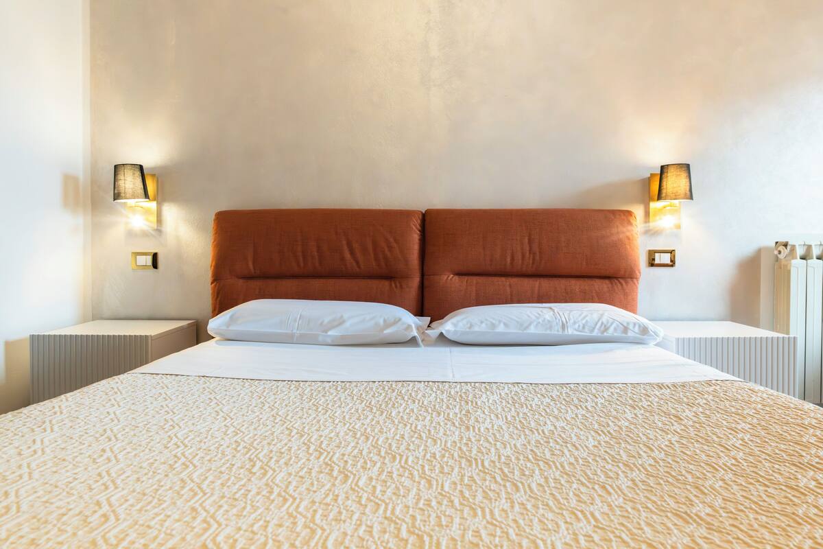 A neatly made bed with white linens and a textured beige blanket, flanked by two bedside lamps with white shades.