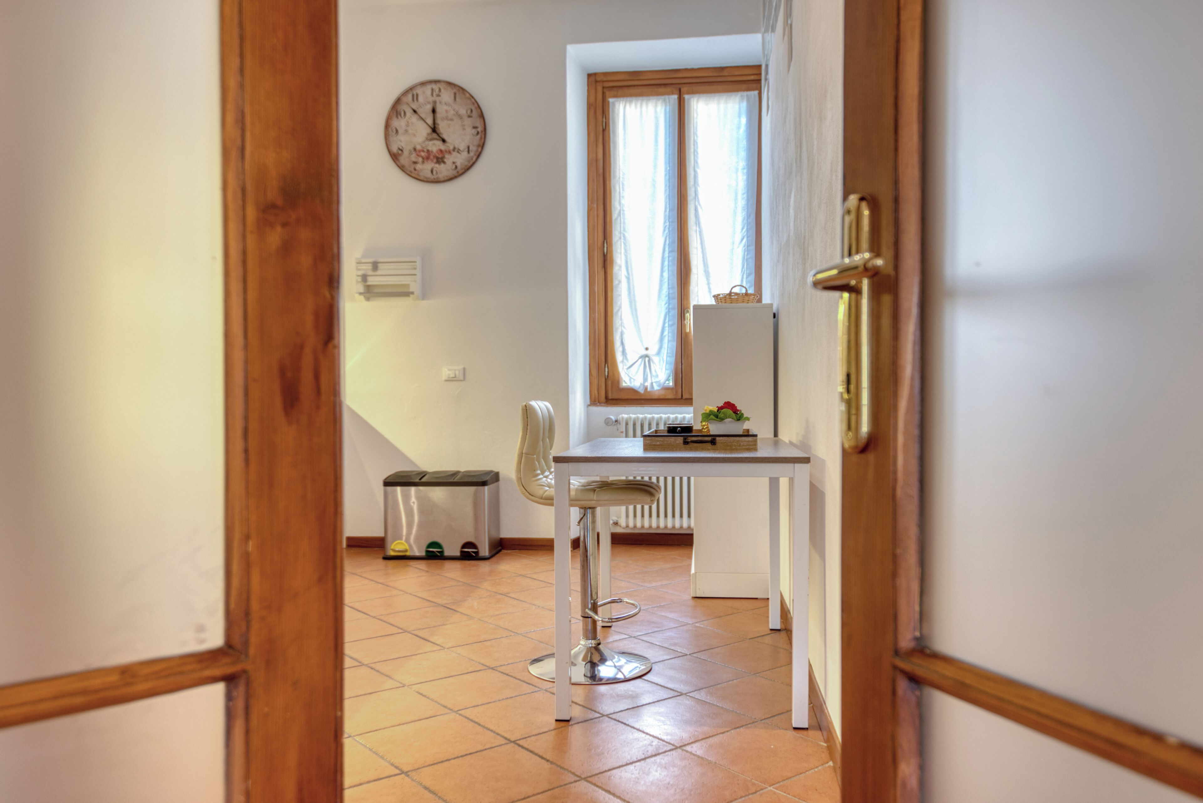 A room with a white table, a chair, a radiator, a clock on the wall, and a window with curtains.