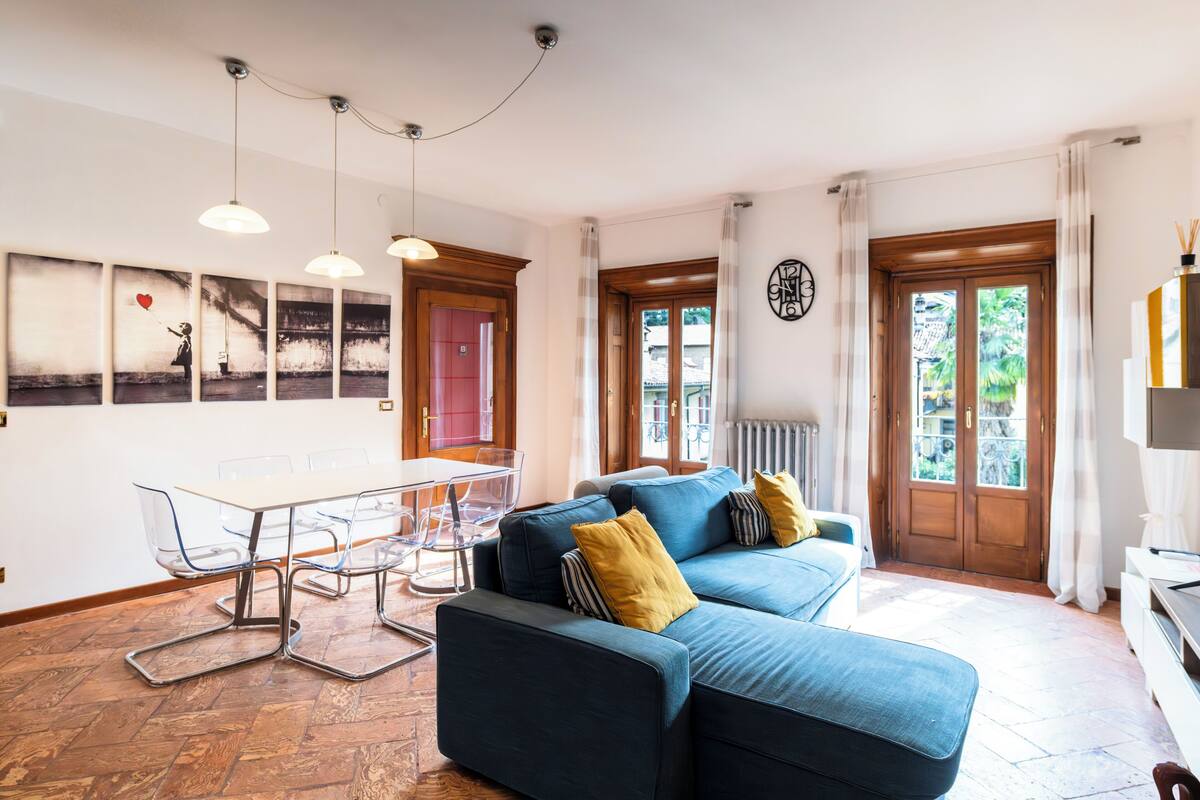 A modern living room with a sofa, a dining table with chairs, and a large window with curtains.