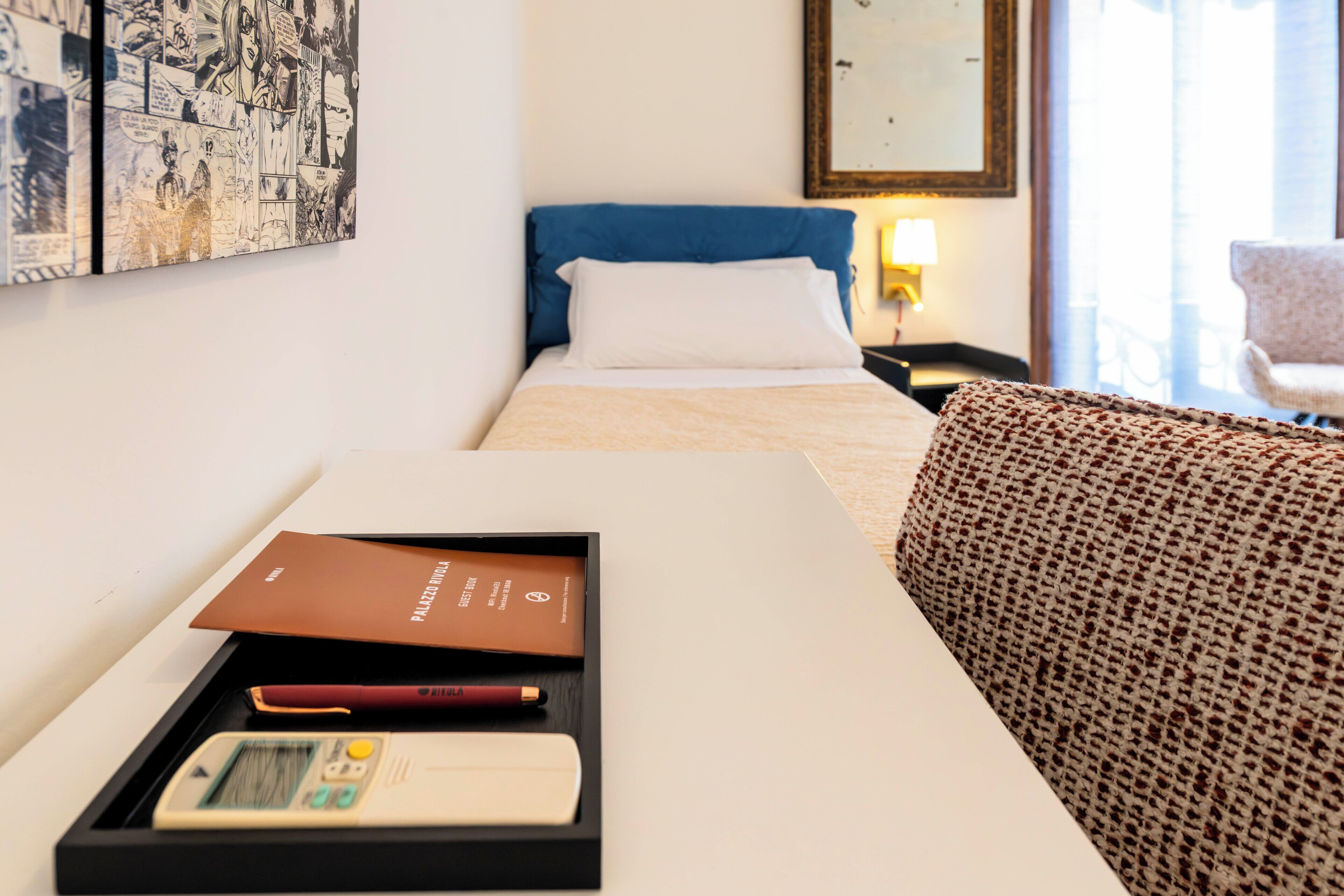 A hotel room with a bed, a bedside table with a book and a remote control, a patterned throw on the couch, and a framed comic strip on the wall.