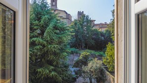 A view through a window of a courtyard with a stone wall, trees, and a building with a bell tower in the background. - Palazzo Rivola (Bergamo)