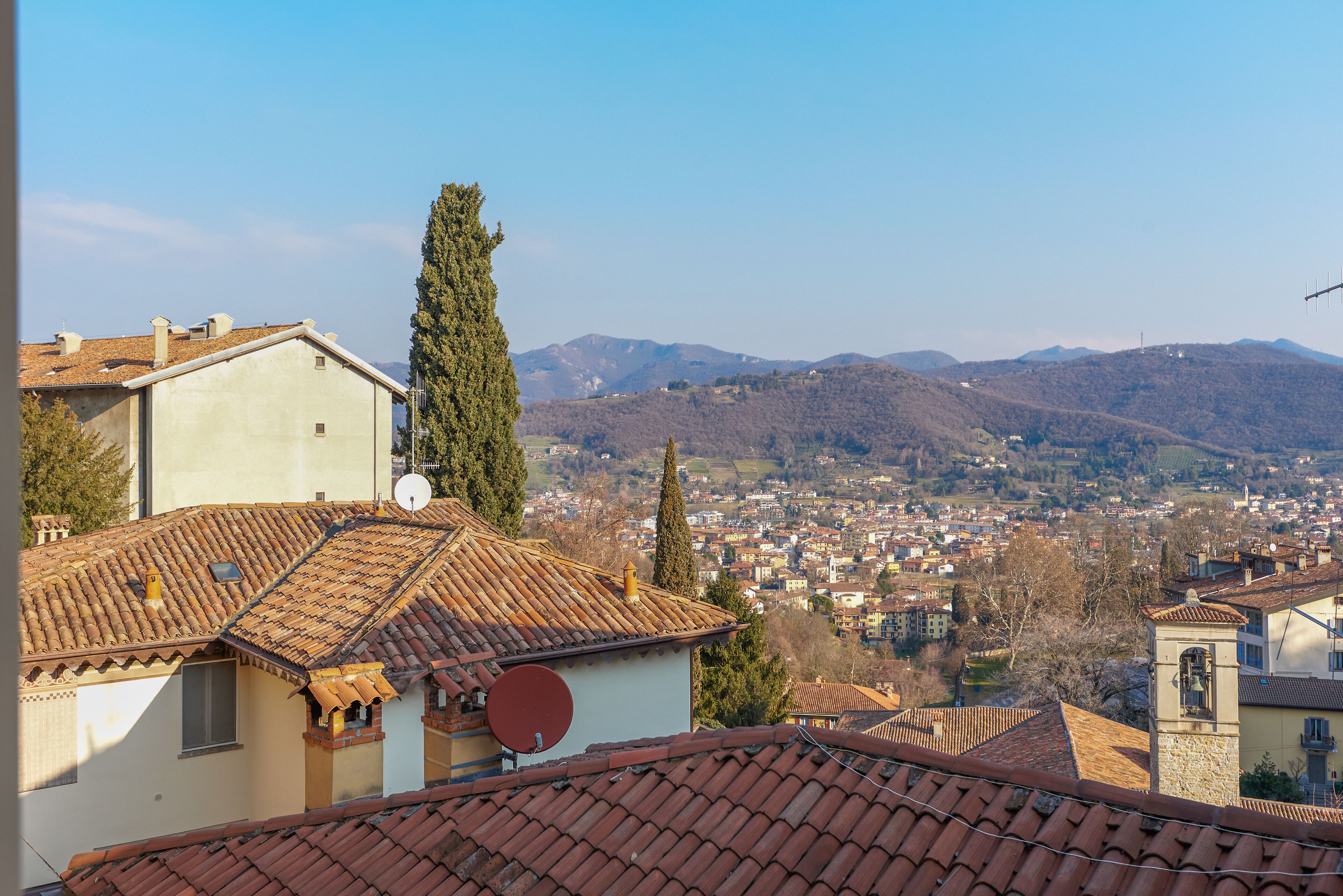 A view of a town with terracotta-tiled roofs, a prominent cypress tree, and distant mountains.