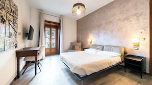 A bedroom with a bed, bedside tables, a desk, a chair, a television, and a door leading to an outdoor area. - Palazzo Rivola (Bergamo)