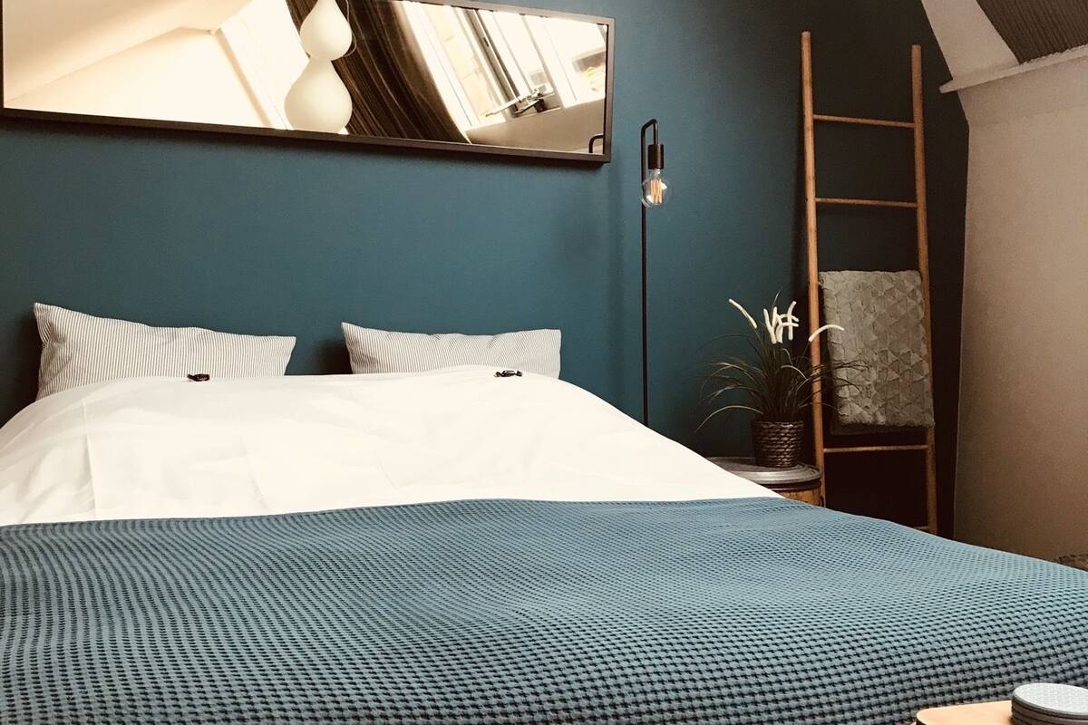 A bedroom with a bed, a bedside table, a mirror, a lamp, and a potted plant.
