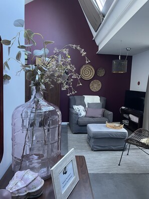 A living room with a large glass vase, a grey sofa, a small ottoman, and a wicker chair. - Guesthouse de Loft (Bruges)