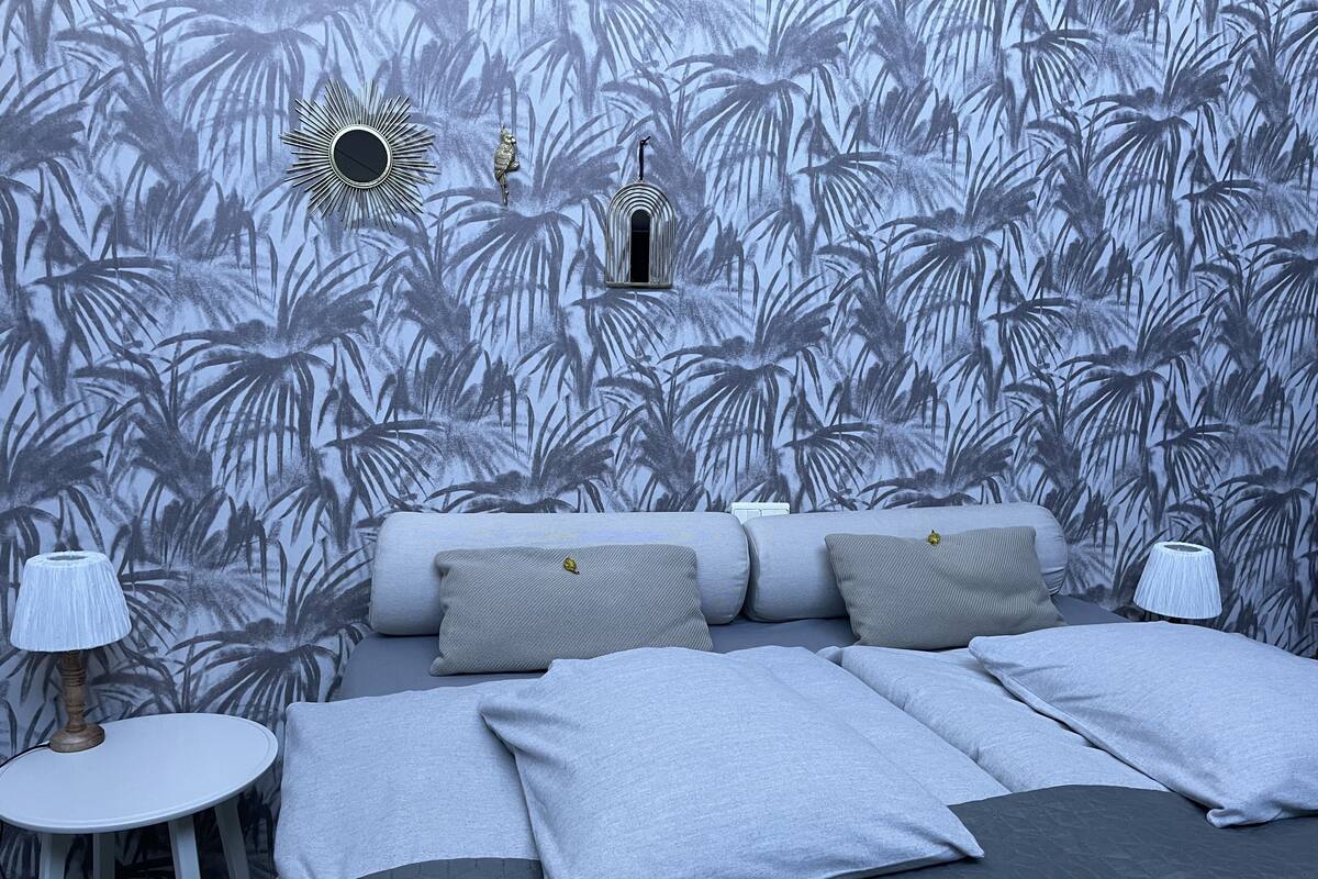 A bedroom with a bed, pillows, a nightstand, a lamp, and a wall with a tropical leaf pattern.