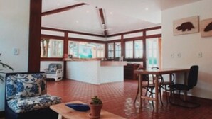 A spacious living area with a sofa, a wooden table, and a dining table with chairs. There is a kitchen area in the background with a window. - Pousada Manacá Parque (Porto Seguro)