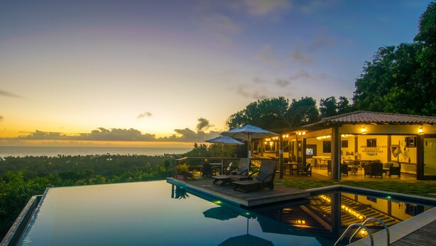 An infinity pool with a wooden deck and lounge chairs, overlooking a scenic view of the ocean at sunset. - Pousada Manacá Parque (Porto Seguro)