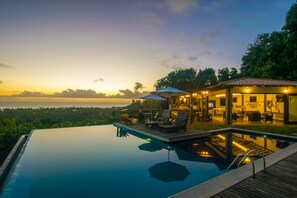 An infinity pool with a wooden deck and lounge chairs, overlooking a scenic view of the ocean at sunset. - Pousada Manacá Parque (Porto Seguro)