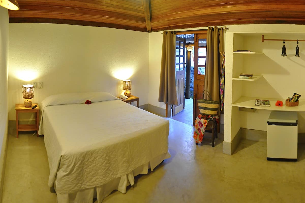 A bedroom with a large bed, bedside lamps, a desk with a chair, and a small refrigerator.