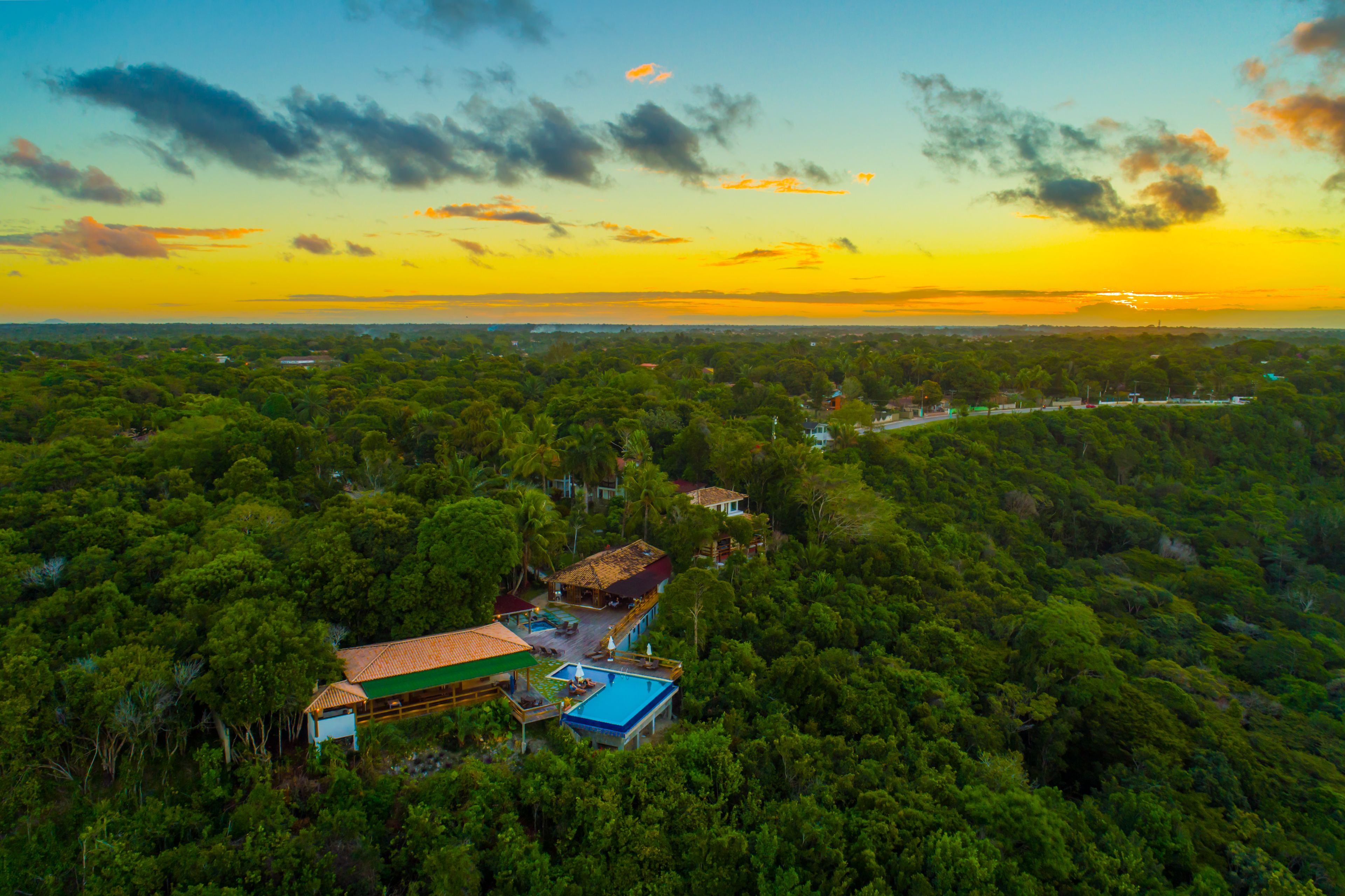 Un complexe hôtelier comprenant plusieurs bâtiments, entouré d’une forêt dense, avec une piscine et une vue sur le coucher de soleil à l’horizon.