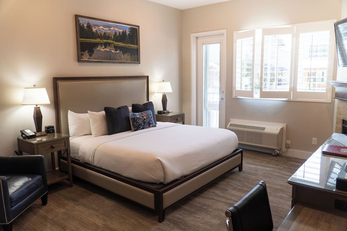 A hotel room with a large bed, two bedside tables with lamps, a desk, and a chair. There is a window with blinds and a painting on the wall.
