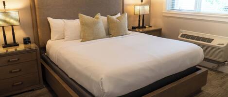 A neatly made bed with a wooden headboard, two bedside tables with lamps, a nightstand, and a window with blinds.