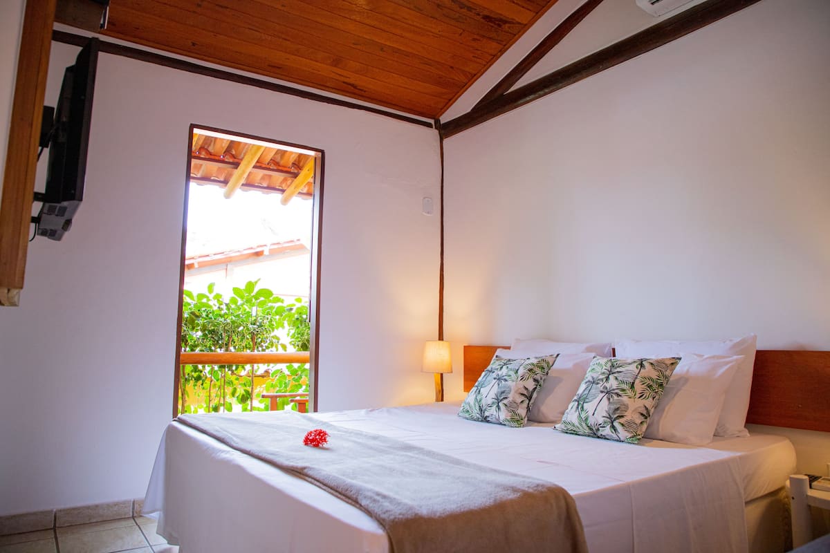 A bedroom with a bed, a nightstand, a window with a view of greenery, and a wooden ceiling.