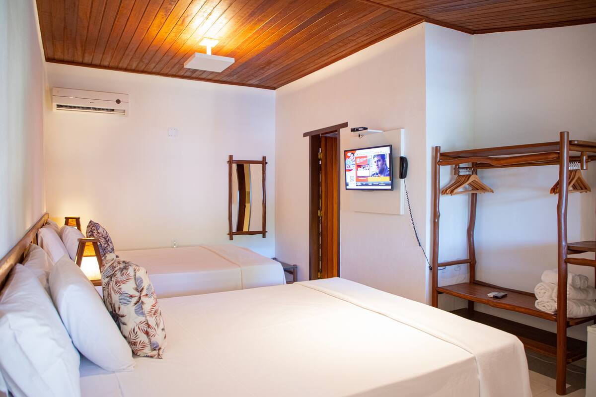 A hotel room with a wooden ceiling, a bed with white linens, a TV mounted on the wall, and a wooden shelf with towels.