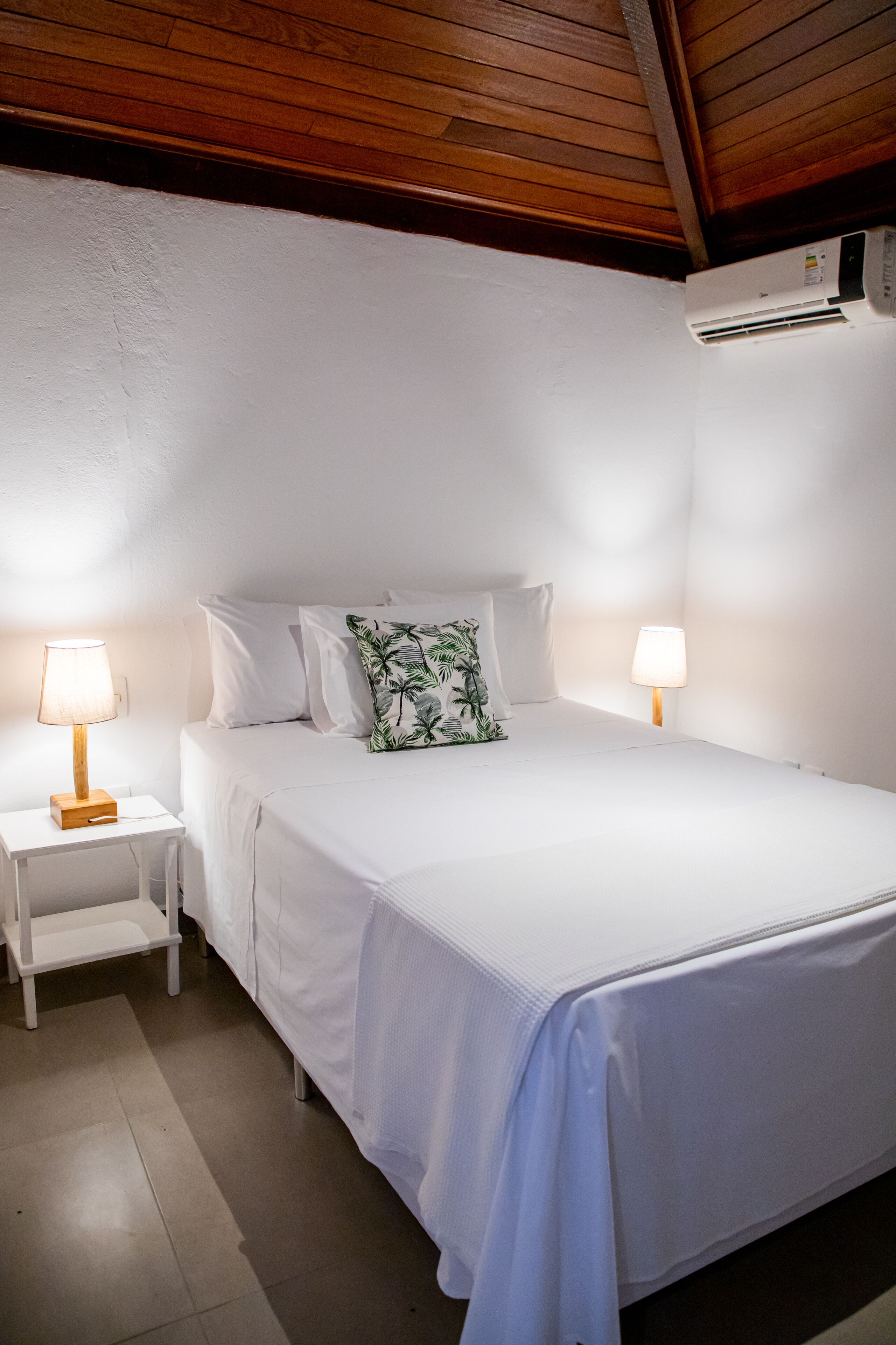 A neatly made bed with white linens and a single green leaf-patterned pillow. There are two bedside tables with lamps, and an air conditioning unit mounted on the wall.