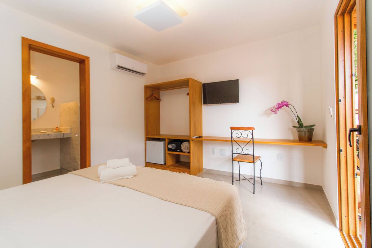 A bedroom with a bed, a wooden wardrobe, a desk with a chair, a potted plant, and a TV on the wall.