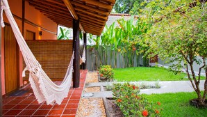 A hammock under a wooden pergola, a small garden with a tree and flowers, and a red-tiled pathway leading to a door. - Pousada Tororão (Porto Seguro)